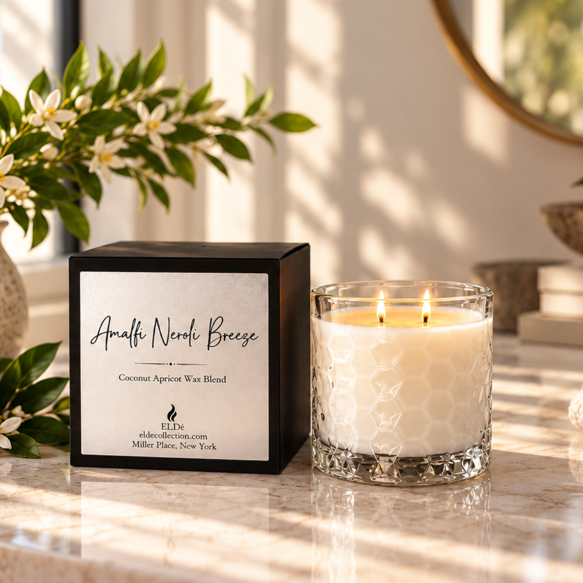 A scented candle in a clear textured glass jar sits on a marble countertop beside its black box packaging. The scene is styled with white flowers in a vase, a sliced lemon, books, and warm sunlight streaming through a window, creating a cozy home interior setting.