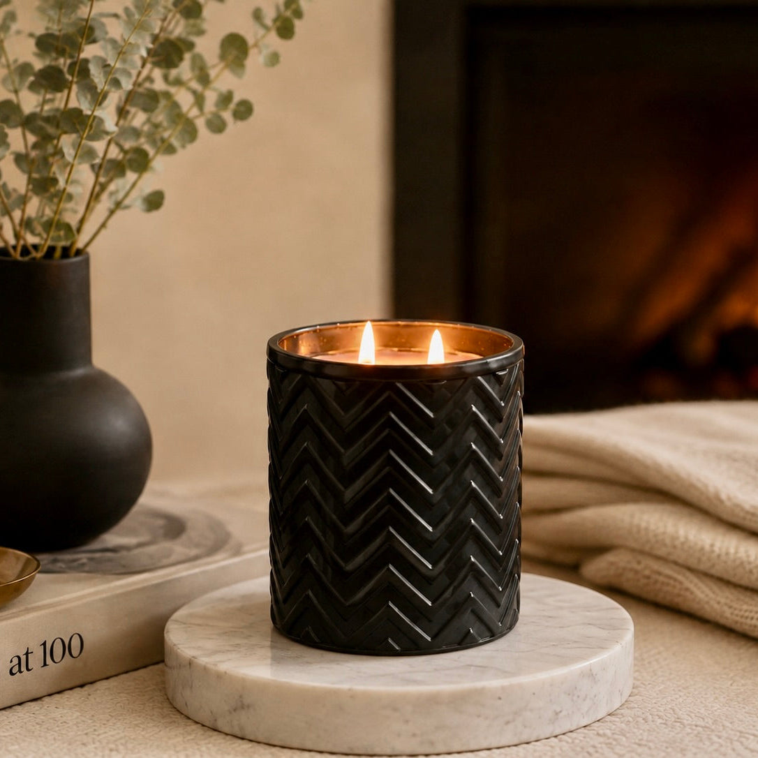 A black textured candle holder with two lit candles sits on a round white marble coaster in a cozy indoor setting. A dark vase with dried leaves, a book, and a folded cream blanket are visible in the background near a blurred fireplace.