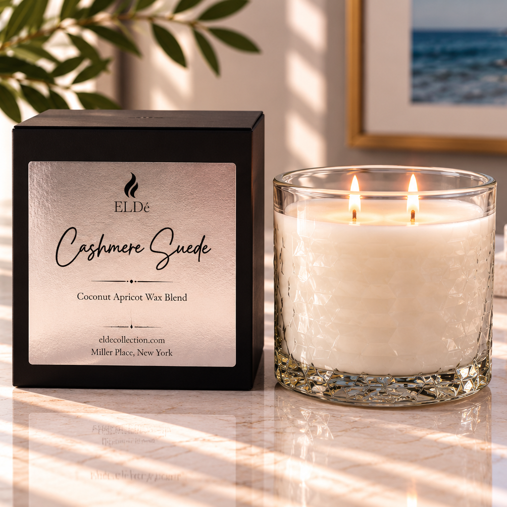A lit double-wick candle in a clear glass jar sits beside its black retail box on a sunlit marble surface. The box label reads ELDE and Cashmere Suede, with coconut apricot win blend and Miller Place, New York visible.