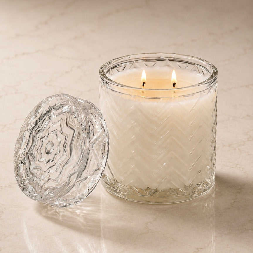 A lit double-wick candle in a clear textured glass jar sits on a light beige marble surface with its matching clear glass lid placed beside it.