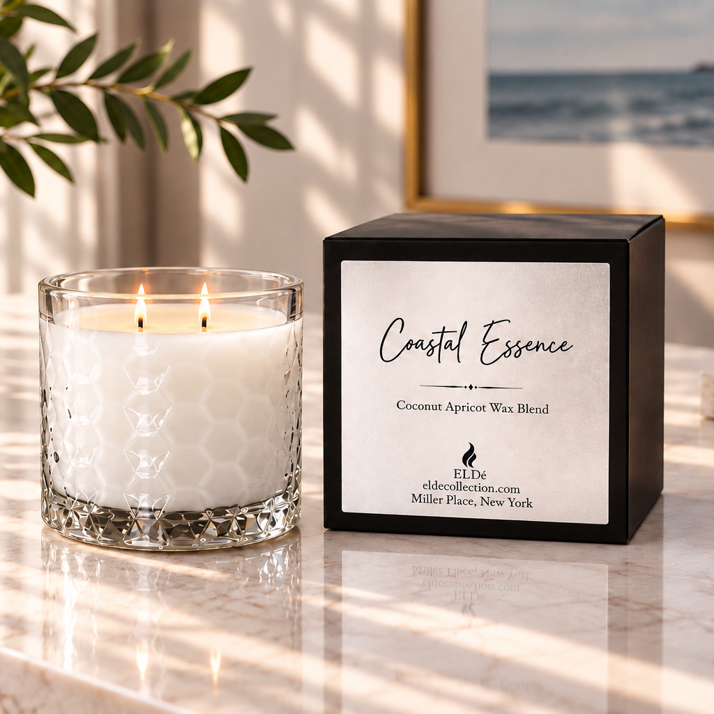 A lit white wax candle in a clear textured glass holder sits beside its black square box labeled 'Coastal Essence' on a marble surface, with a potted plant and framed ocean artwork in the background.