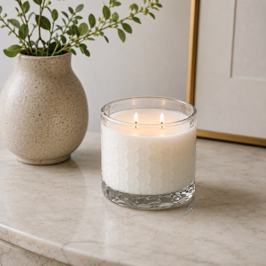 A lit white candle in a clear textured glass jar sits on a light marble surface, with a speckled beige vase of greenery and part of a framed artwork in the background.