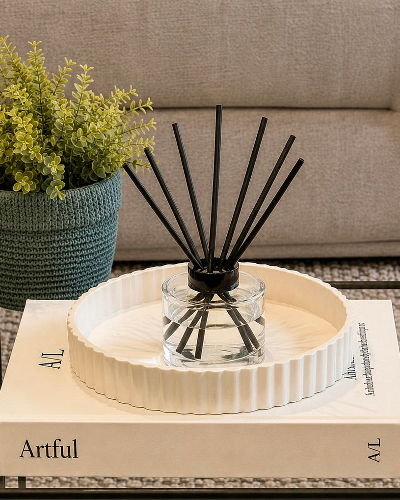 A reed diffuser in a clear glass bottle with black reeds, set in a white ribbed ceramic tray on top of a cream book on a glass table. A green potted plant sits in the background next to a beige sofa.