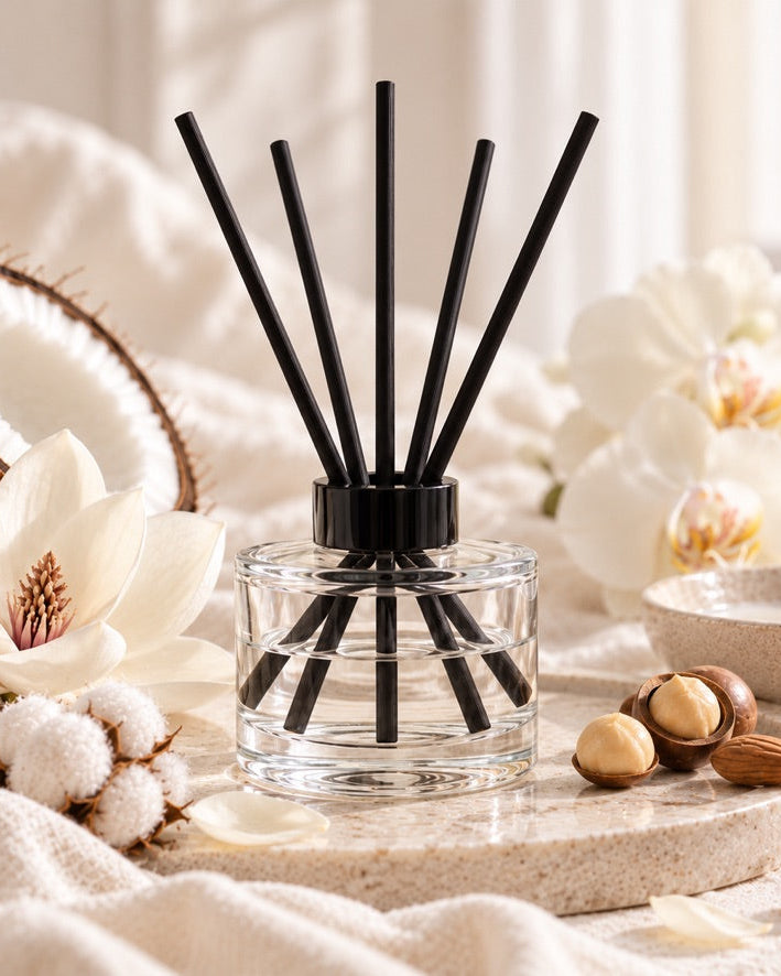 A clear glass reed diffuser bottle with black reeds and a black cap, styled on a stone tray with white flowers, cotton bolls, almonds, and a coconut in a soft neutral setting.