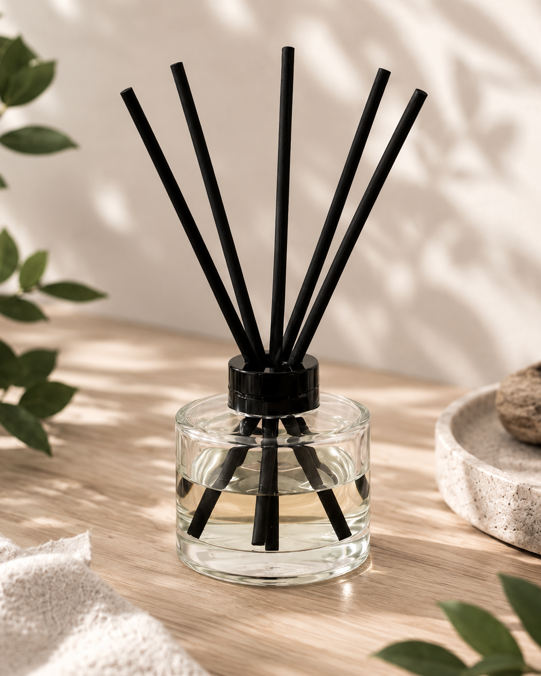 A clear glass reed diffuser bottle with a black cap and multiple black reeds, placed on a light wood surface with soft shadows, green leaves, and a textured stone dish in the background.