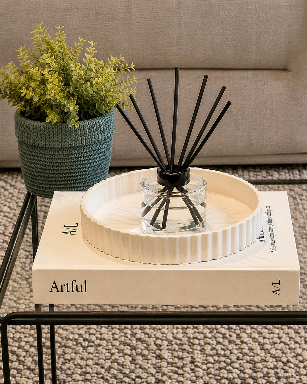 A reed diffuser in a clear glass bottle with black reeds, set in a white ribbed ceramic tray on top of a cream book on a glass table. A green potted plant sits in the background next to a beige sofa.