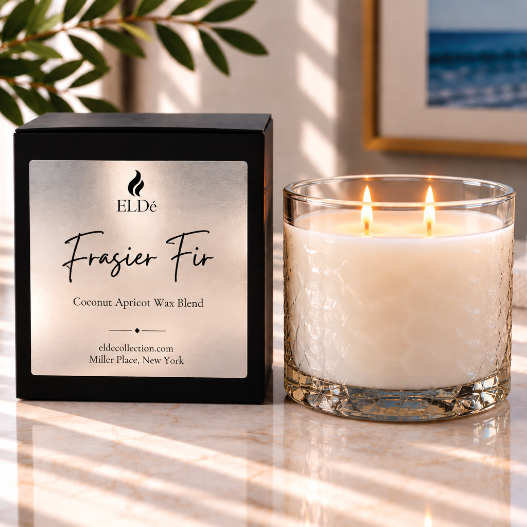 A lit scented candle in a clear glass vessel sits beside its black retail box on a sunlit marble surface. The box label reads ELDE and Frasier Fir, with coconut apricot wax blend text below.
