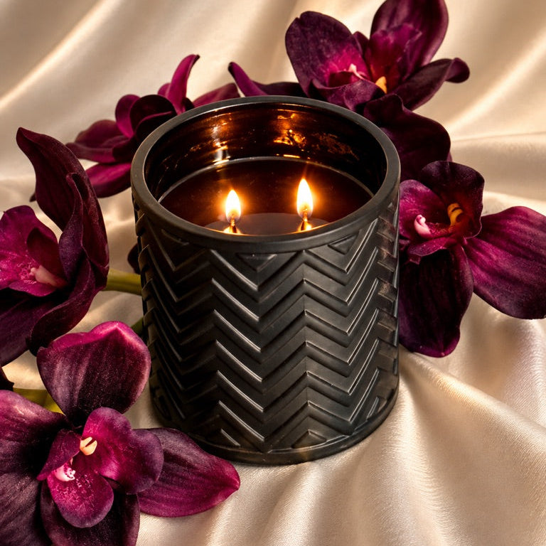 A black textured candle holder with two lit candles, styled with deep purple orchids on a draped champagne satin fabric background.