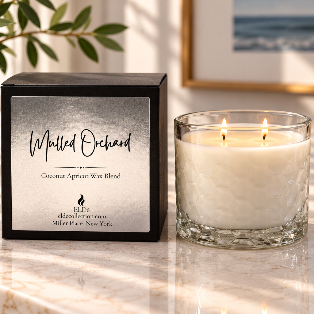 A lit double-wick candle in a textured clear glass jar sits beside its black retail box on a marble surface. The label reads 'Mulled Orchard' and 'Coconut Apricot Wax Blend,' with the brand ELDE visible near the bottom.