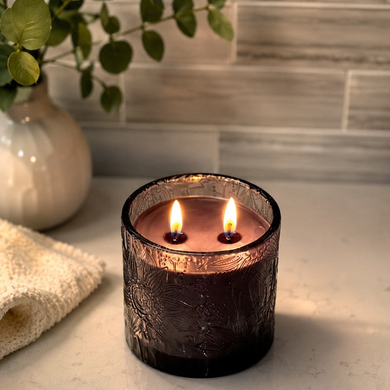 A lit two-wick candle in a dark amber textured glass jar sits on a light marble countertop with a folded cream towel and a vase of green leaves in the background.