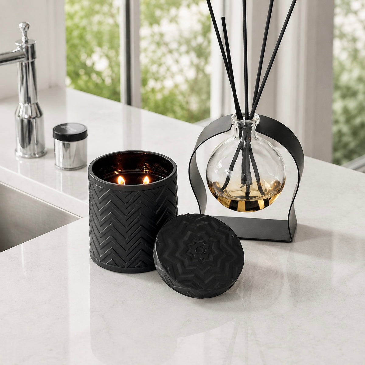 A black textured candle in a ribbed cylindrical holder with a lit flame, shown beside a matching black reed diffuser and glass bottle on a white marble countertop near a sink.