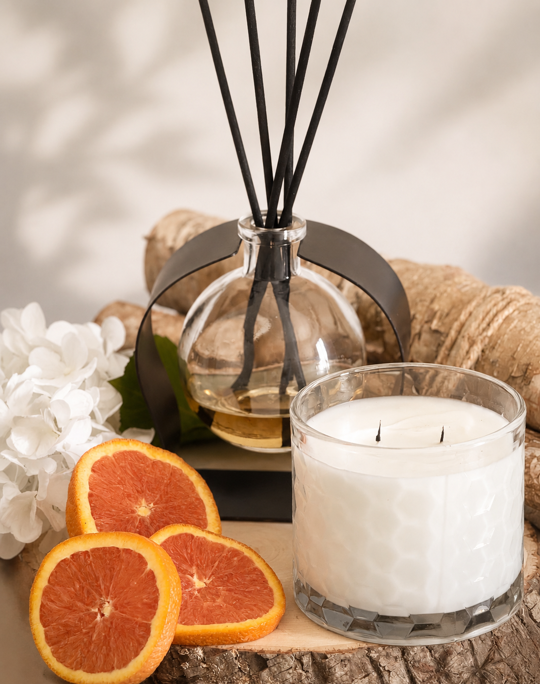 A styled product shot featuring a clear glass reed diffuser bottle with black reeds and a black collar, alongside a white wax candle in a faceted clear glass jar. The scene is decorated with three sliced orange wedges, white flowers, and natural wood textures against a softly lit neutral background.