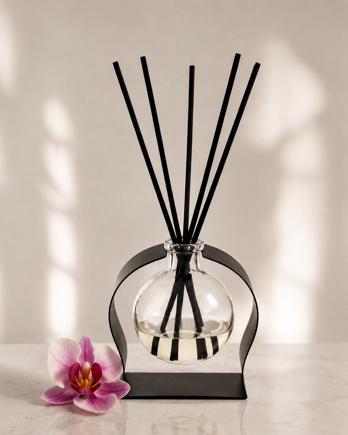 A reed diffuser with a clear glass round bottle, black metal stand, and six black reeds, styled on a light marble surface with a pink orchid flower and soft window shadow lighting.