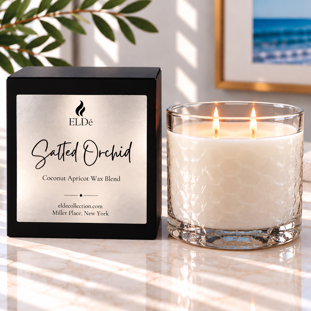 A lit candle in a clear textured glass jar sits beside its black rectangular box on a bright marble surface. The box label reads 'ELDe Salted Orchid Coconut Apricot Wax Blend.