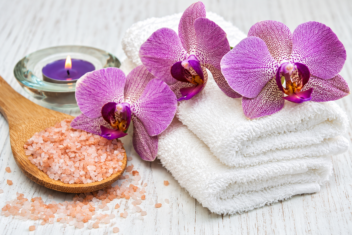 Three white towels, three purple orchid flowers, pink salt crystals in a wooden spoon, and a small lit purple candle on a white wooden surface.