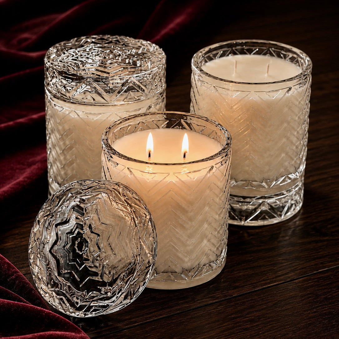 Three clear cut-glass candle holders with white wax candles on a dark wooden surface, with one lid placed in front and deep burgundy velvet fabric in the background.