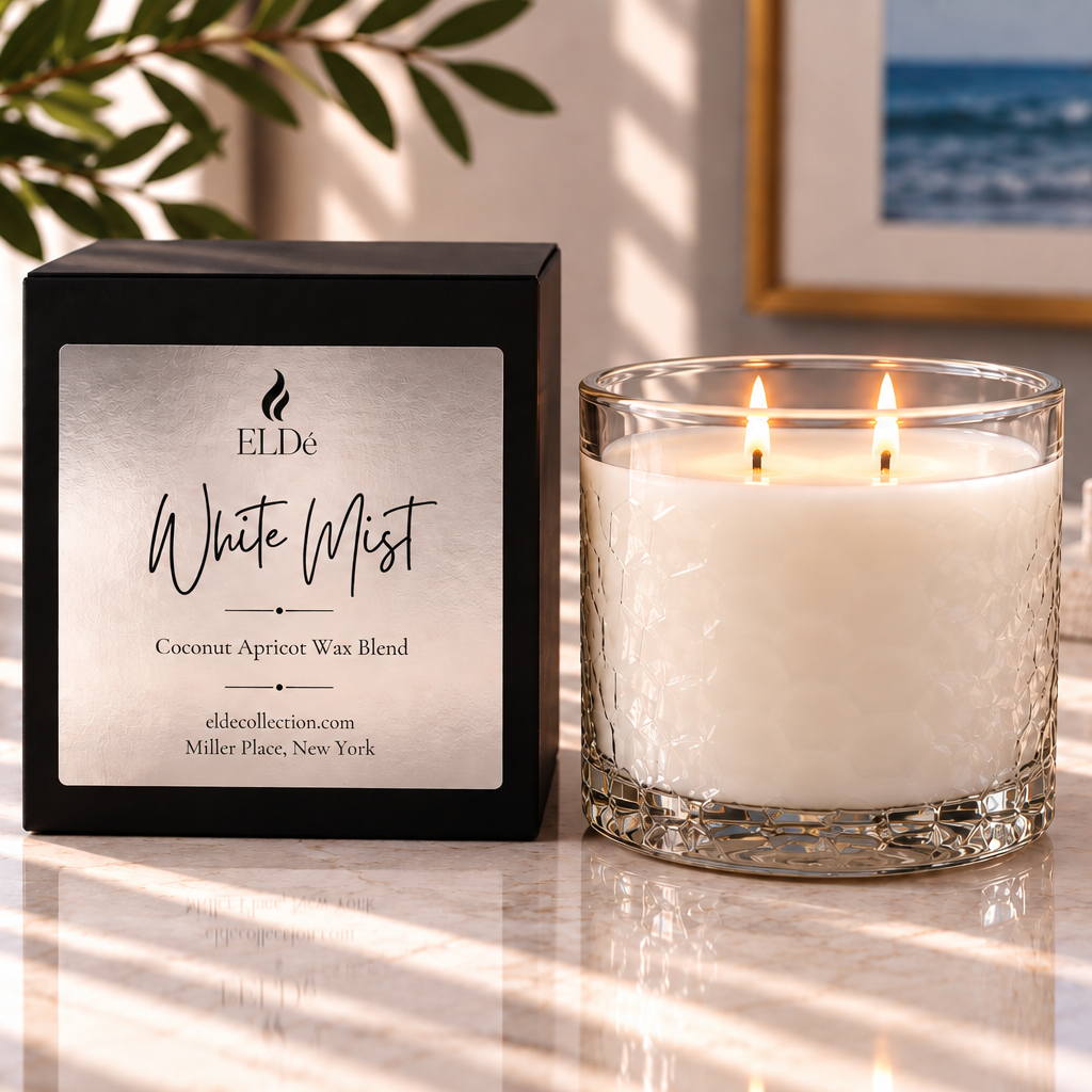 A lit white candle in a clear textured glass jar sits beside its black square box on a sunlit marble surface. The box label reads ELDE and Coconut Apricot Wax Blend.