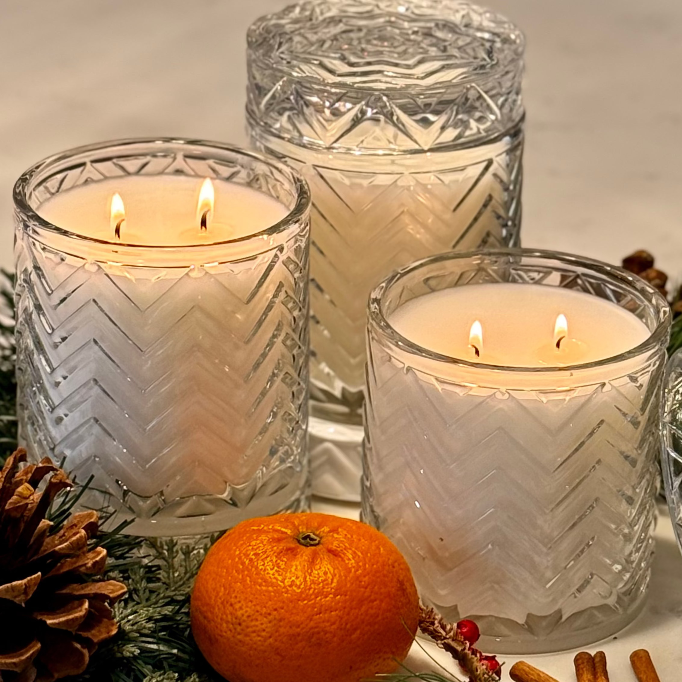 Three lit candles in decorative glass holders with an orange and pinecones on a table.