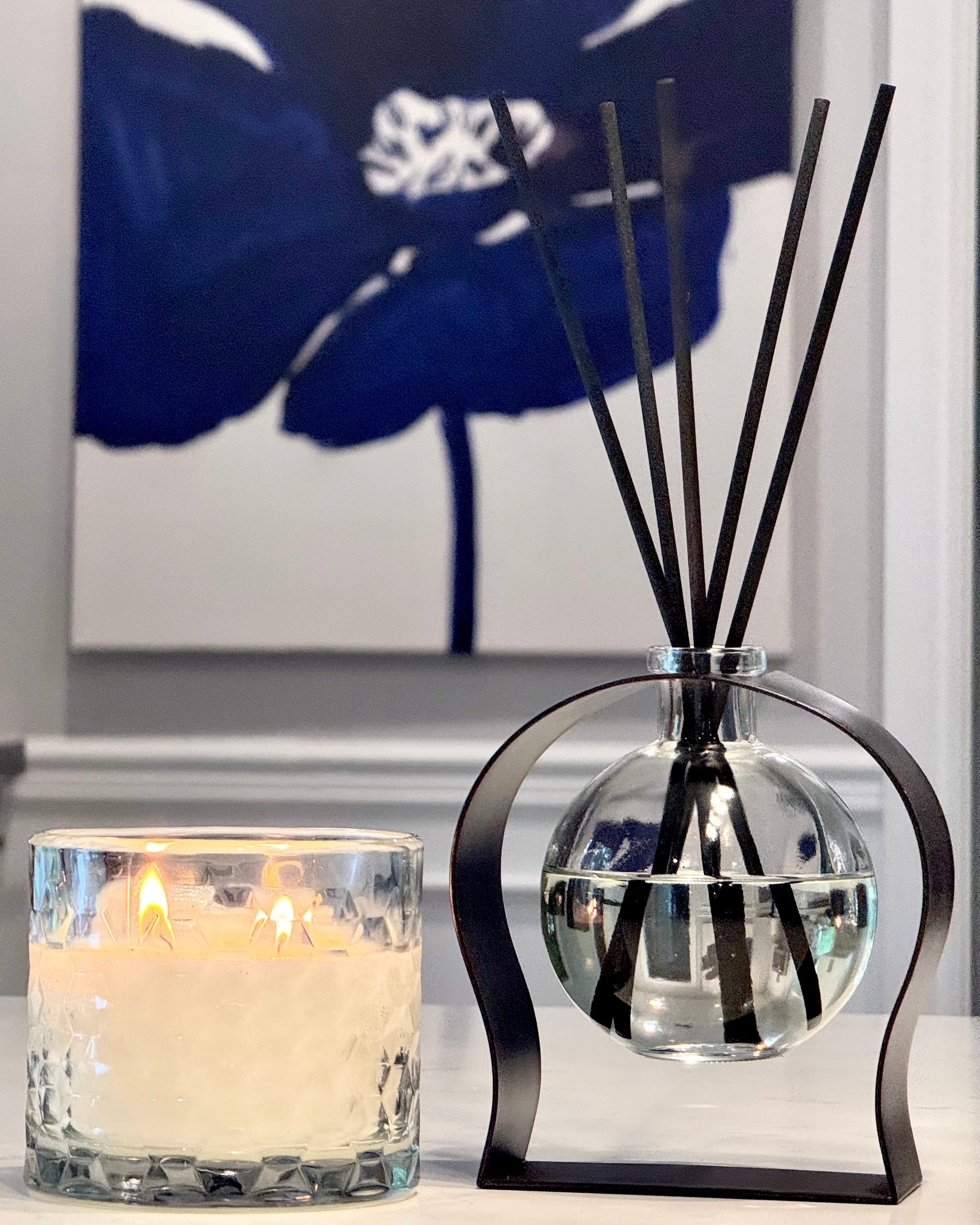 Diffuser with black sticks and a candle in front of a blue floral painting