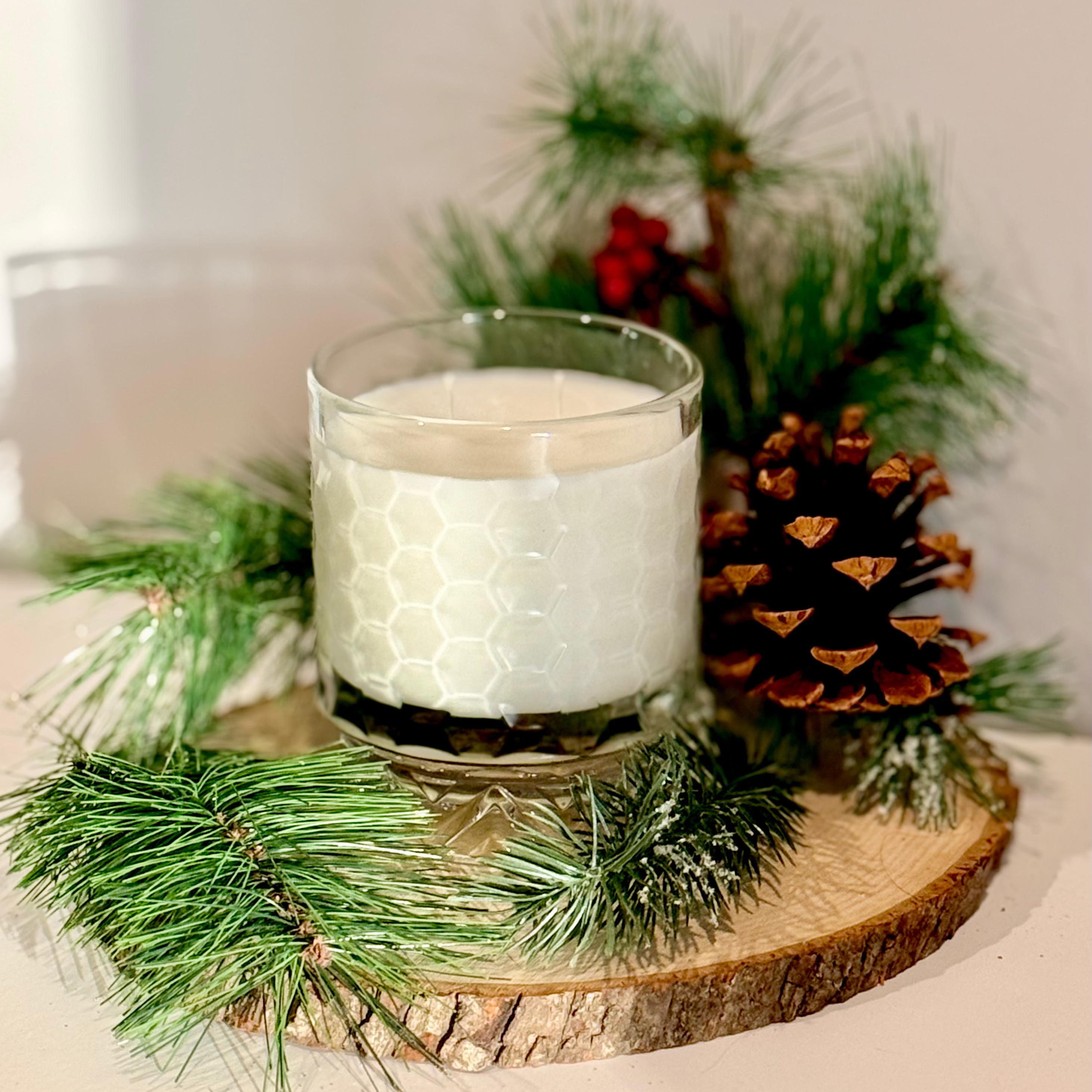 White candle in a glass holder on a wooden base with pine branches and a pine cone.