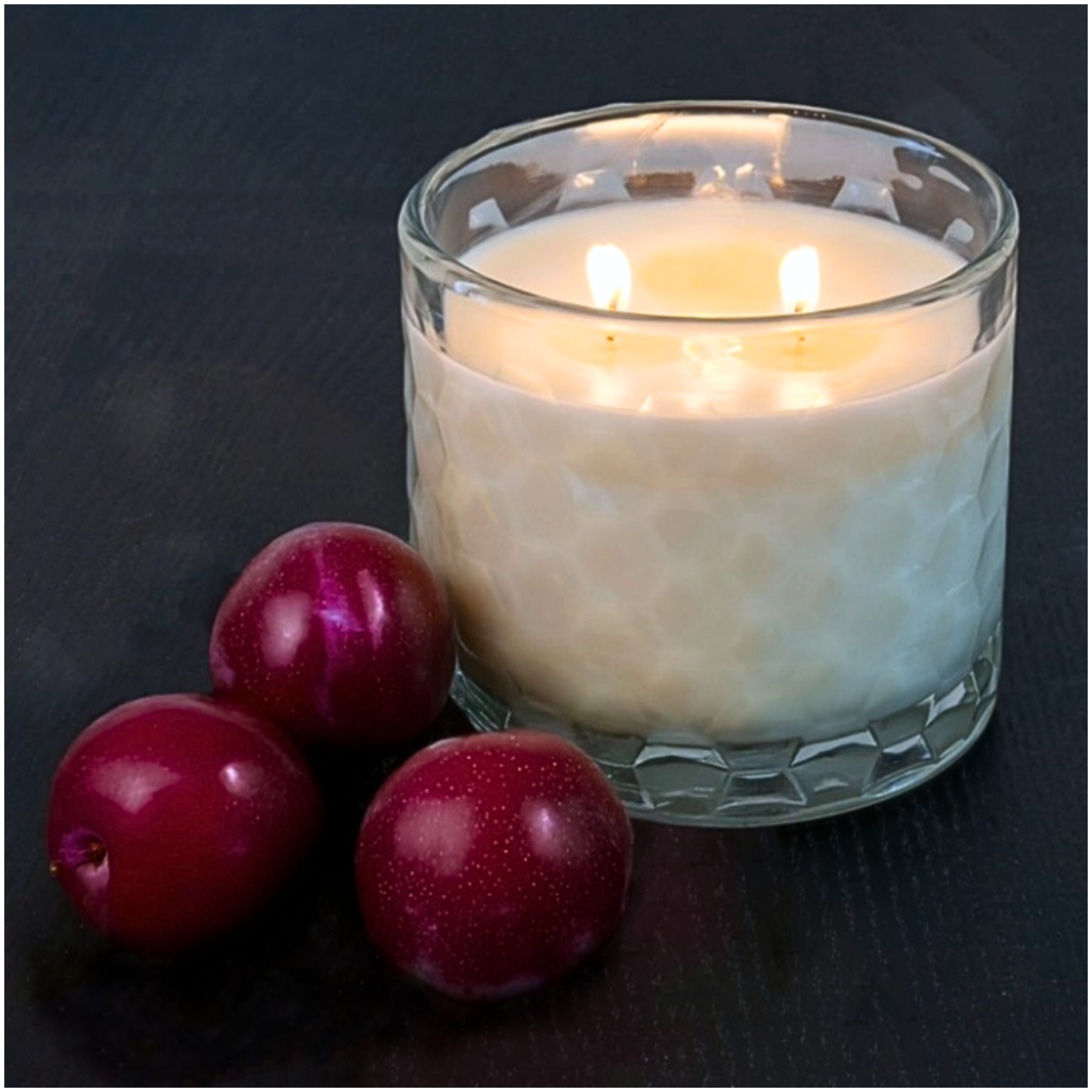 Candle in a glass jar with three plums on a dark surface