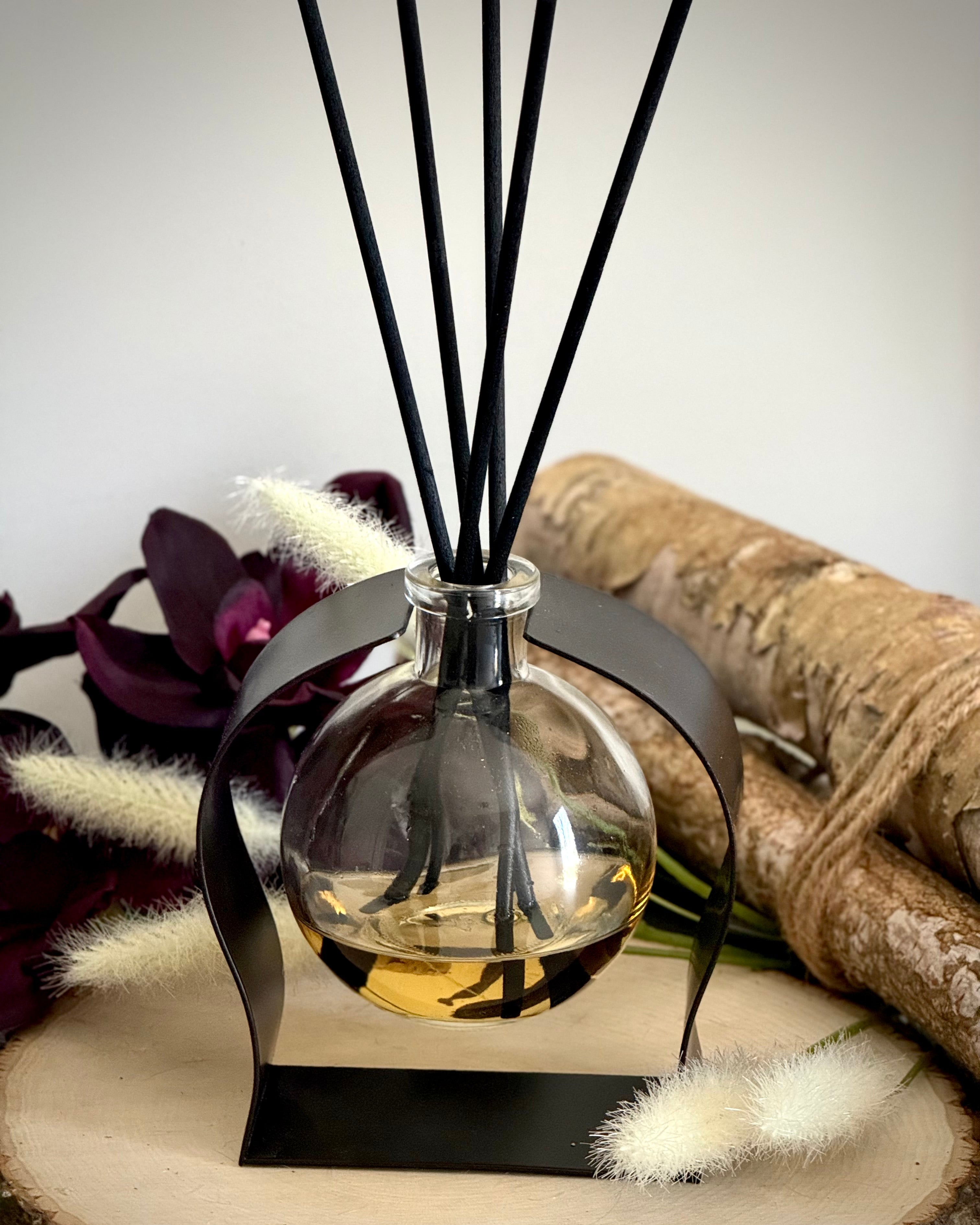 Aromatherapy diffuser with black sticks on a wooden surface with flowers and fabric in the background.