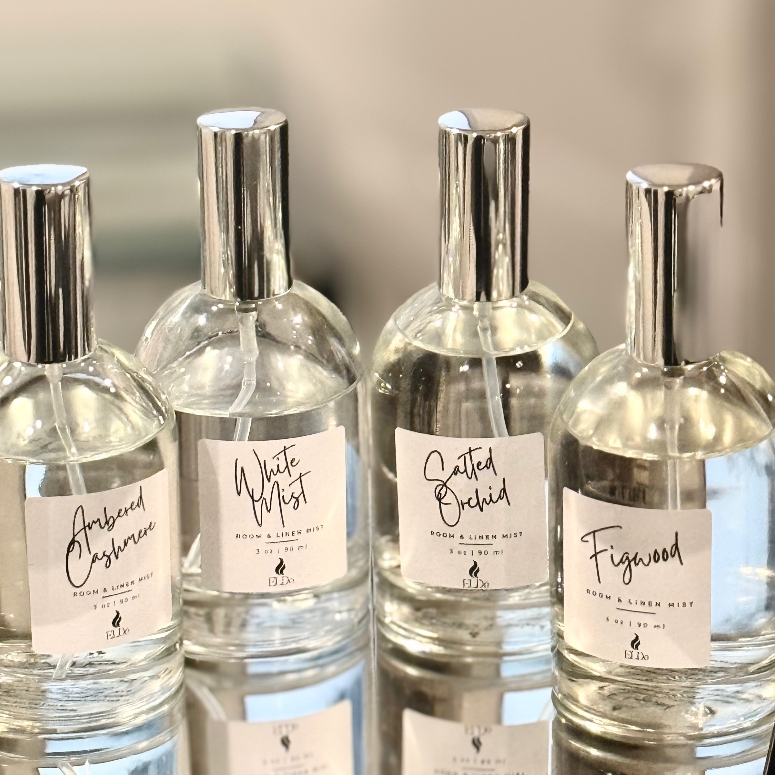 Four clear perfume bottles with silver caps and labels on a reflective surface.