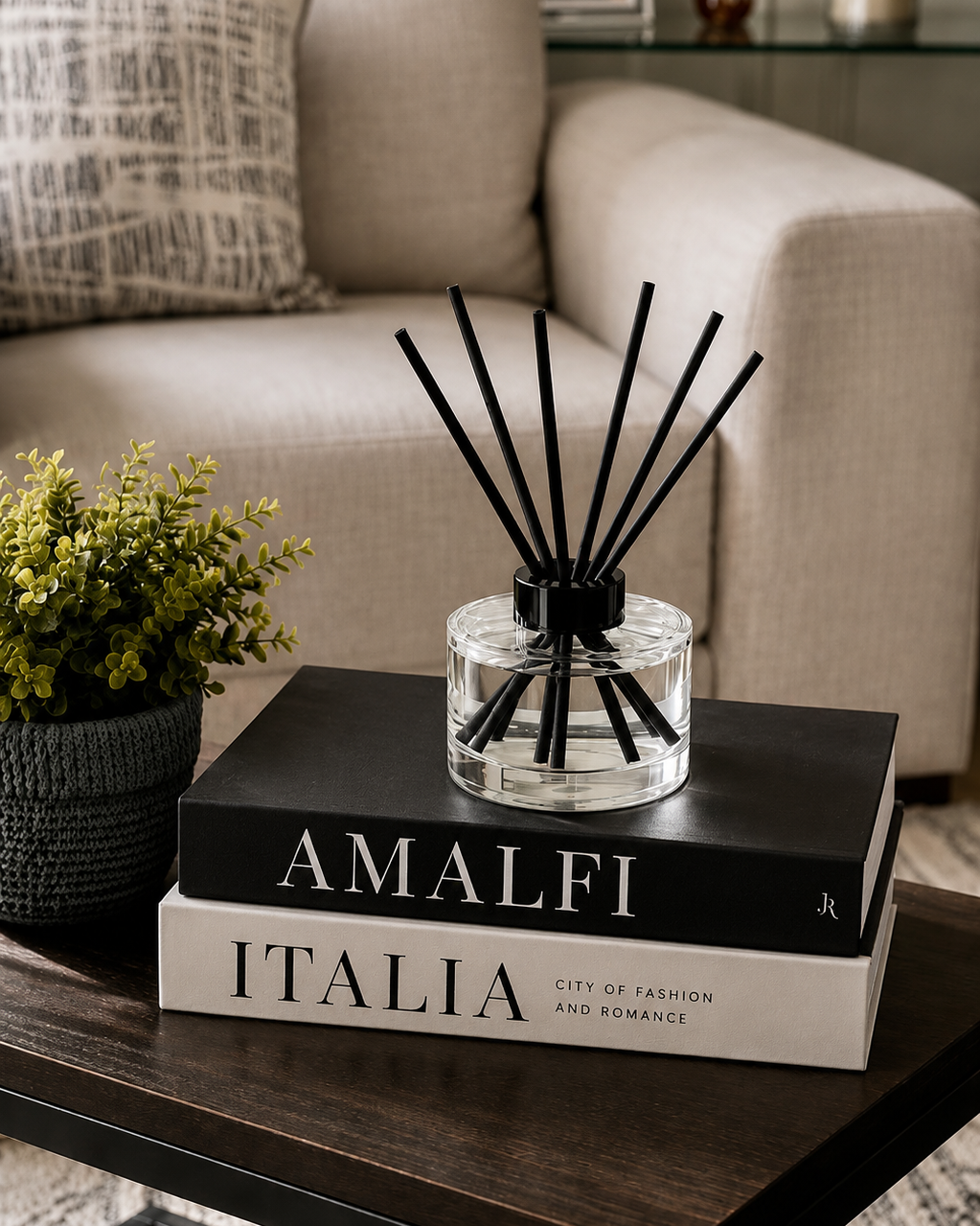 A reed diffuser with black reeds in a clear glass bottle sits on top of two stacked books on a dark wooden table in a living room setting.