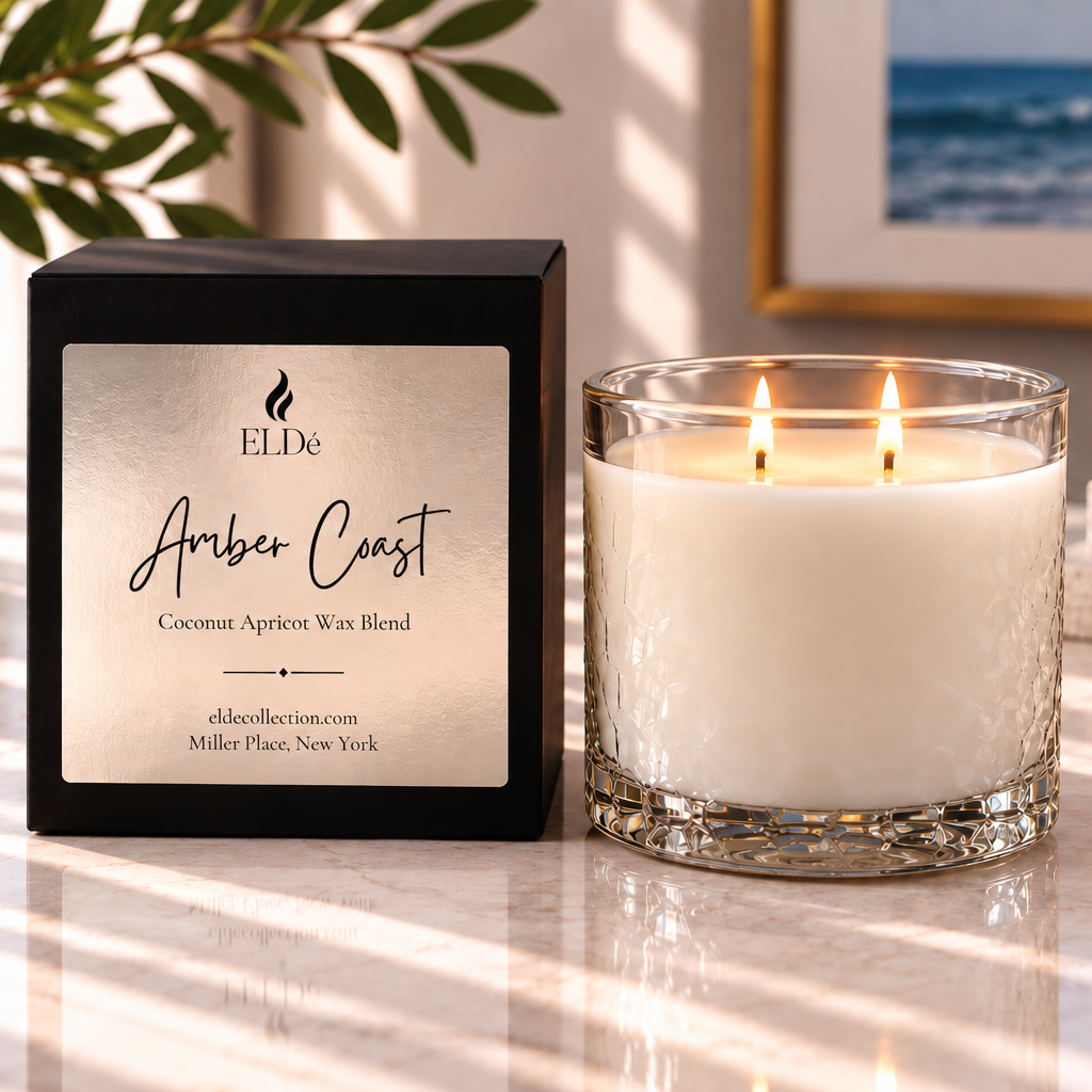 A double-wick scented candle in a clear textured glass jar sits beside its black branded box on a marble surface. The candle wax is white, and the box label reads ELDe Amber Coast Coconut Apricot Wax Blend.