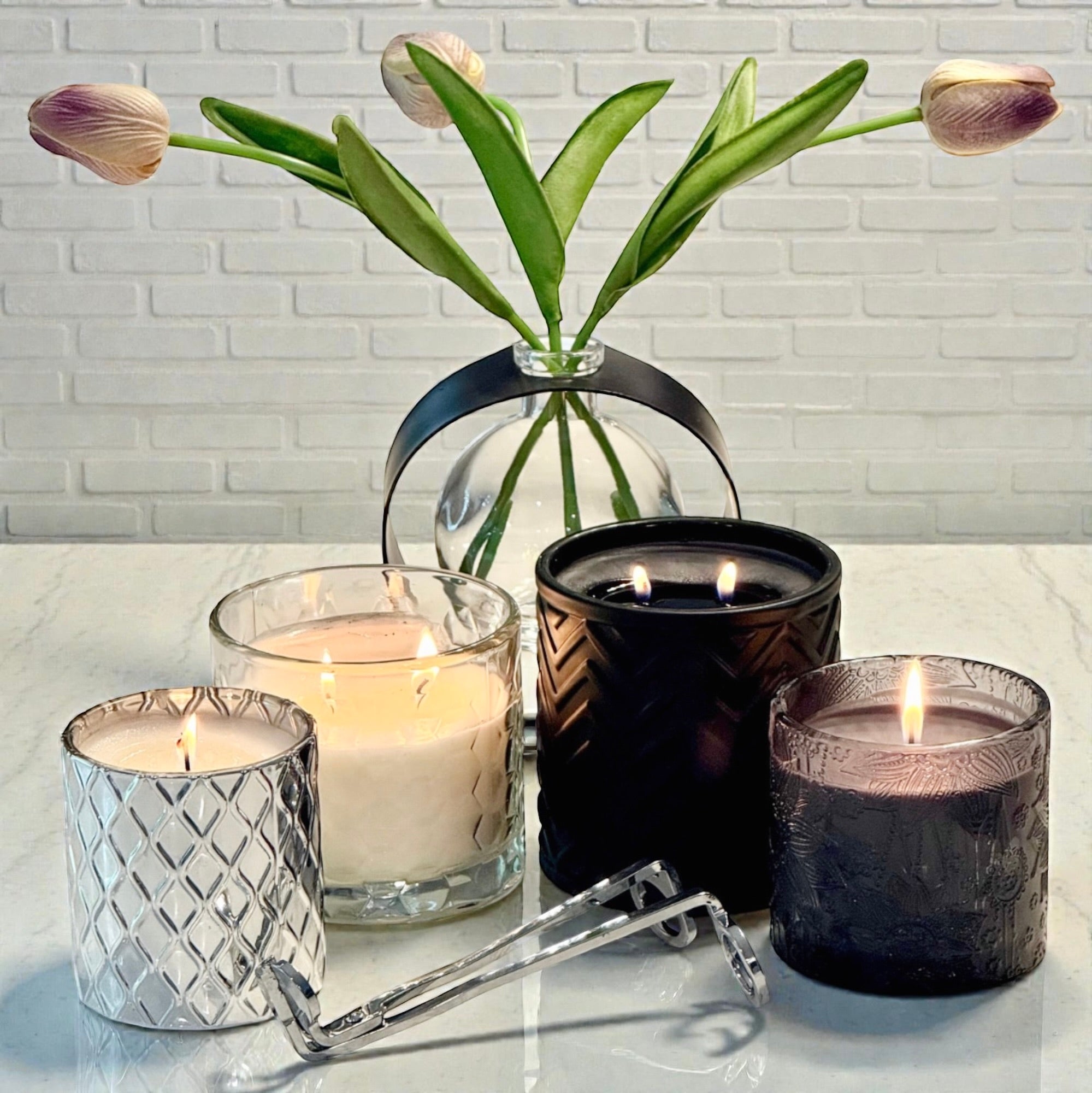 Four candles in different holders with a vase of tulips on a white surface.