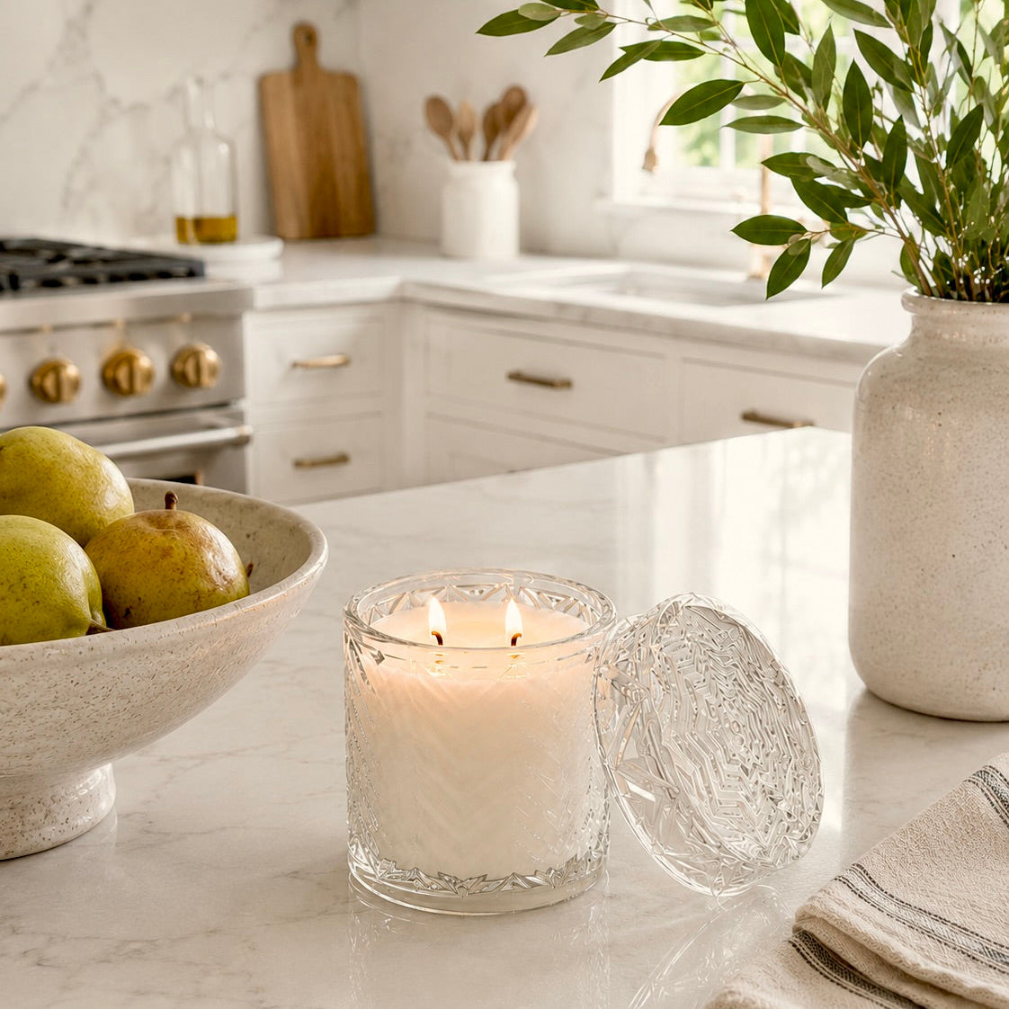 A lit white candle in a clear textured glass jar sits on a white marble kitchen counter with its matching glass lid leaning beside it. A bowl of pears, a vase with greenery, and a modern kitchen are softly blurred in the background.
