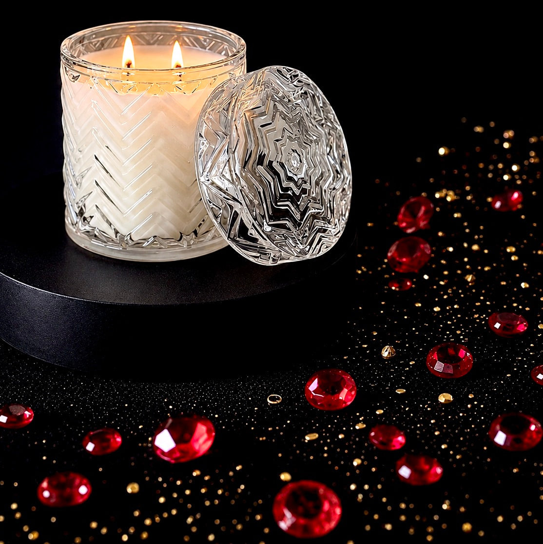 A lit white candle in a clear textured glass jar sits beside a matching clear faceted glass lid on a black pedestal, with scattered red gem-like beads and gold confetti on a black background.