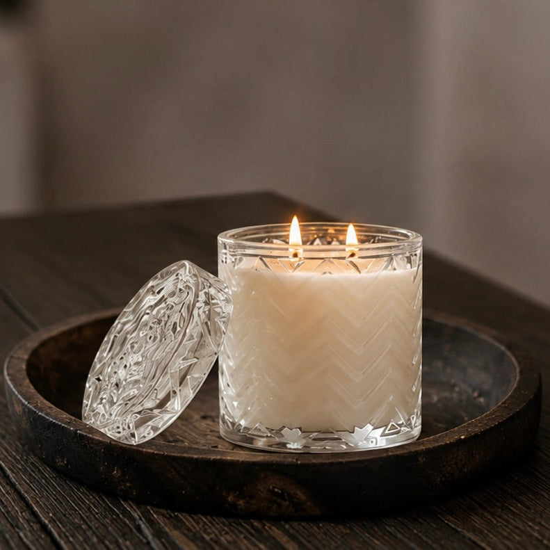 A lit double-wick candle in a clear textured glass jar sits on a dark round wooden tray, with a matching clear glass lid resting beside it on a dark wood tabletop.