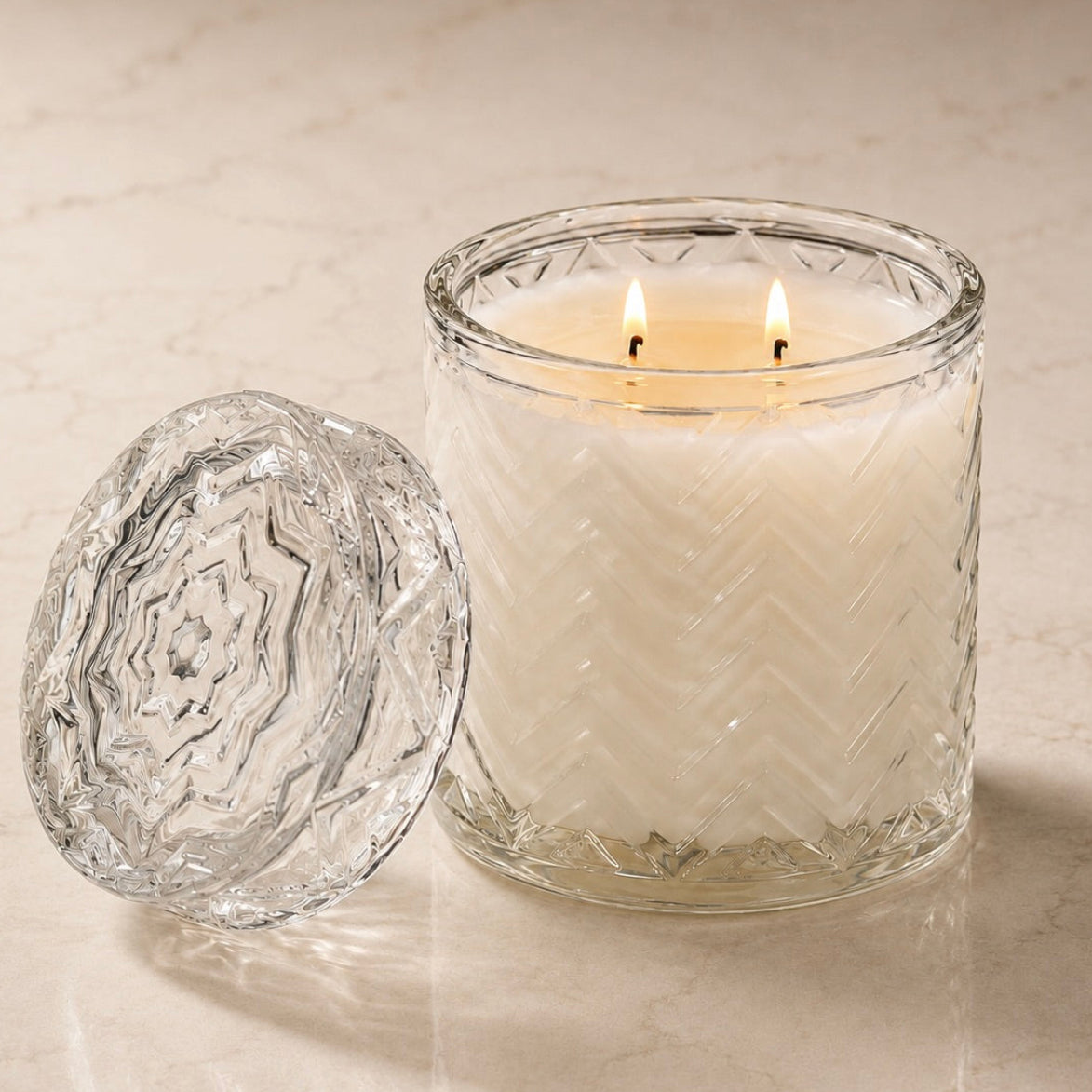A lit double-wick candle in a clear textured glass jar sits on a light beige marble surface with its matching clear glass lid placed beside it.