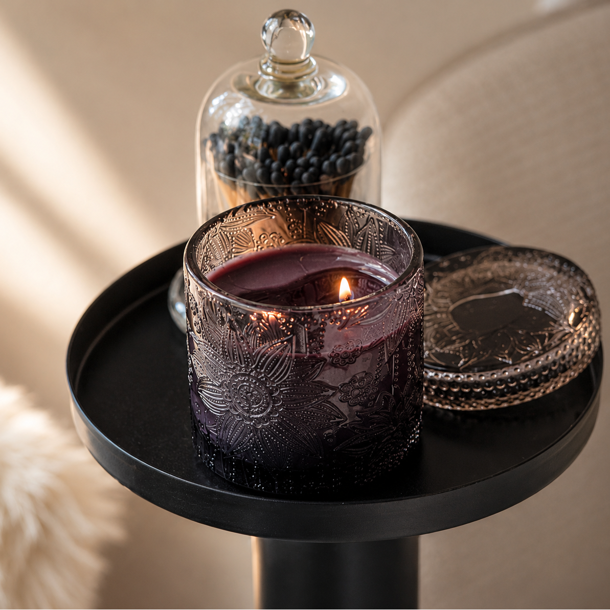 A lit candle in an etched dark purple glass jar sits on a black side table, with its matching lid beside it. A glass cloche filled with dark berries and a beige upholstered chair are visible in the softly lit background.