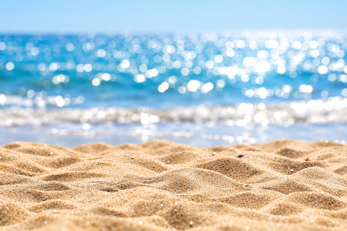 A close-up view of golden sand in the foreground with a blurred ocean background showing bright blue water and sparkling sunlight.