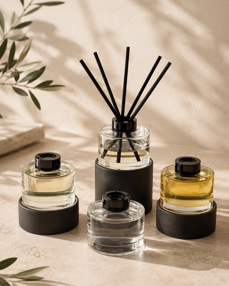 Four reed diffusers with black caps and black reeds, arranged on a beige surface with soft shadows and green leaves in the background.