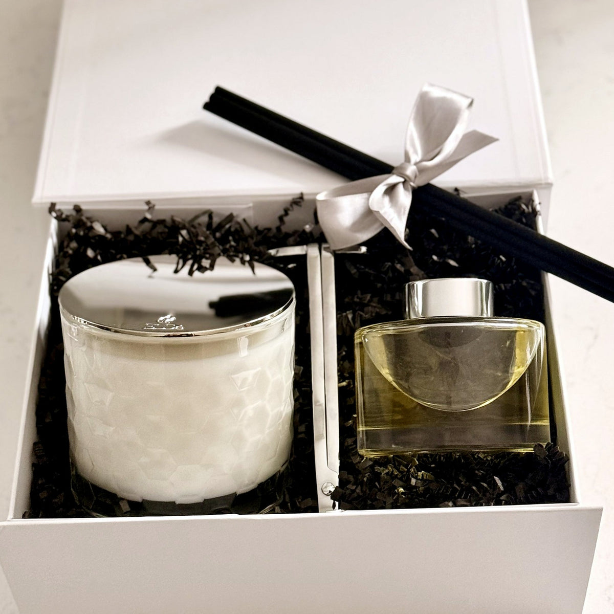 A gift set in a white box containing a white candle with a silver lid and a clear glass diffuser with liquid, and a silver wick trimmer all resting on black shredded paper. A silver ribbon bow and  black reed diffuser sticks are placed on top.