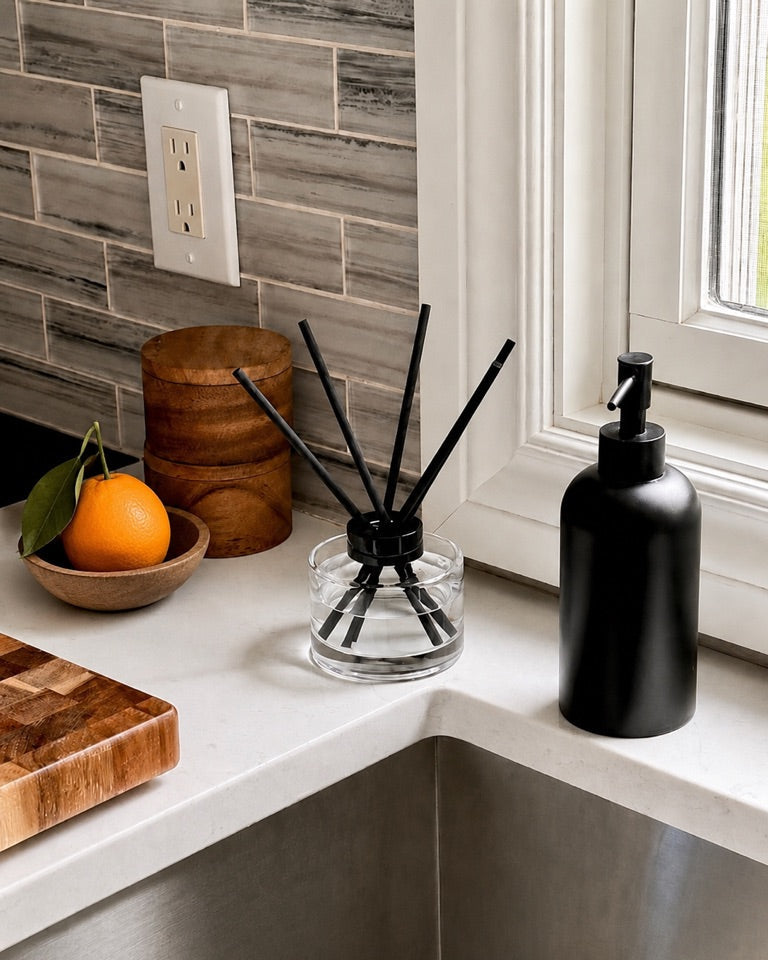 A matte black pump bottle and a clear glass reed diffuser with black reeds sit on a white kitchen countertop beside a window, with a tiled backsplash, wooden bowl, and orange in the background.
