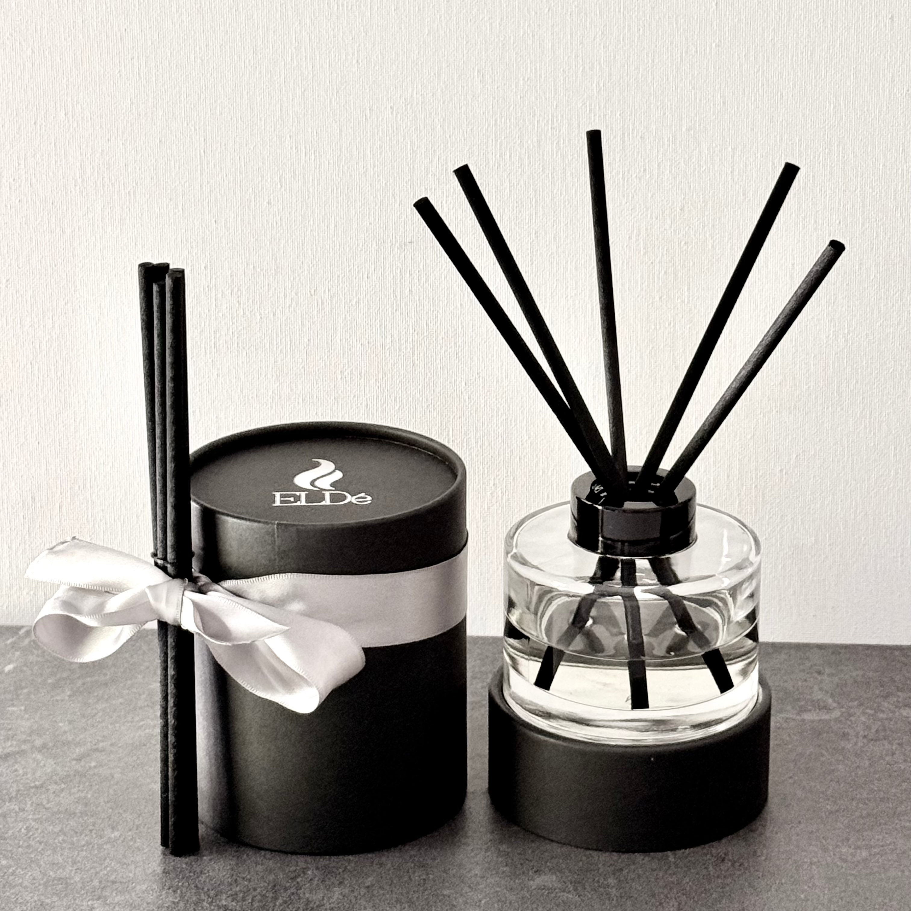 Two diffusers, one in a black container with a white bow and the other in a clear container with black sticks, on a gray surface.