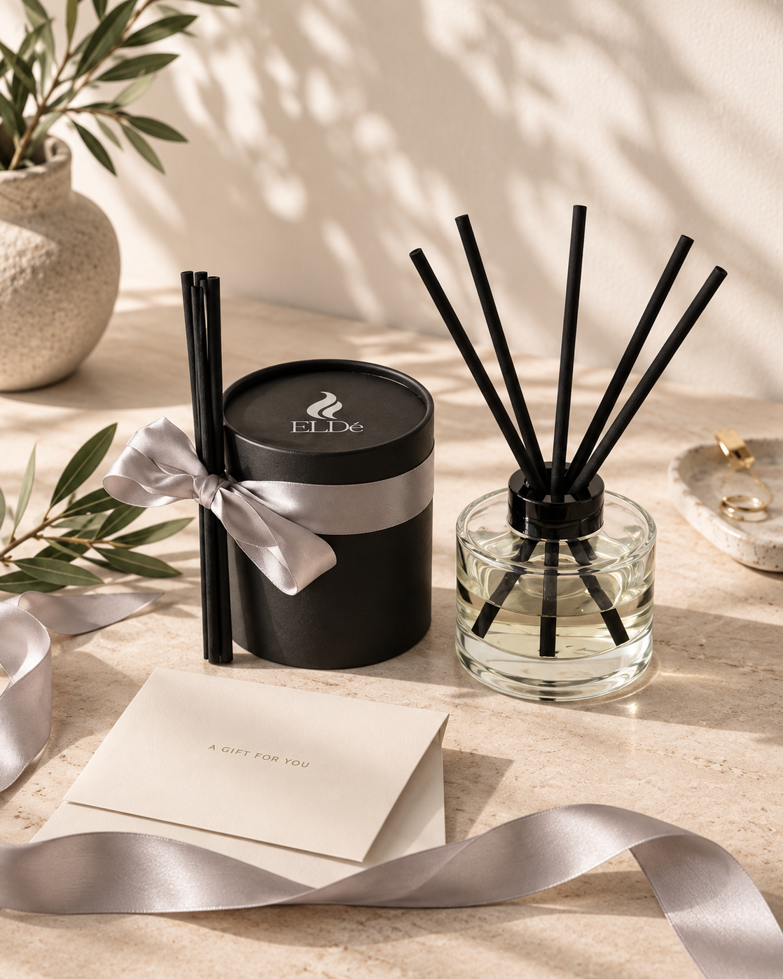A reed diffuser set on a warm beige stone surface, featuring a black cylindrical tin with a silver ribbon and lid printed with 'ELLE', alongside a clear glass bottle with black reeds. A cream gift card reading 'A GIFT FOR YOU' and a silver ribbon are arranged nearby, with soft leaf shadows cast across the scene.