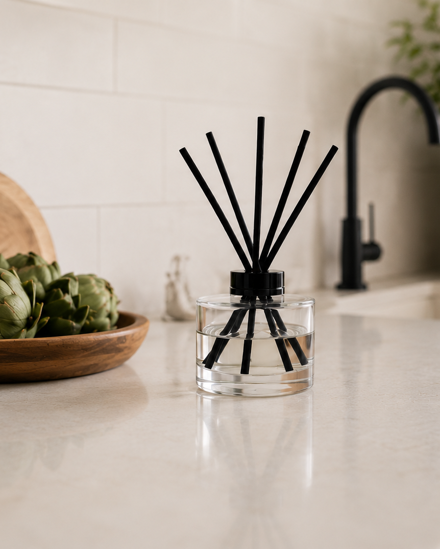 A clear glass reed diffuser bottle with black reeds stands on a light stone countertop in a bright kitchen setting. A bowl of artichokes, a wooden cutting board, a black faucet, and a potted plant are visible in the background.