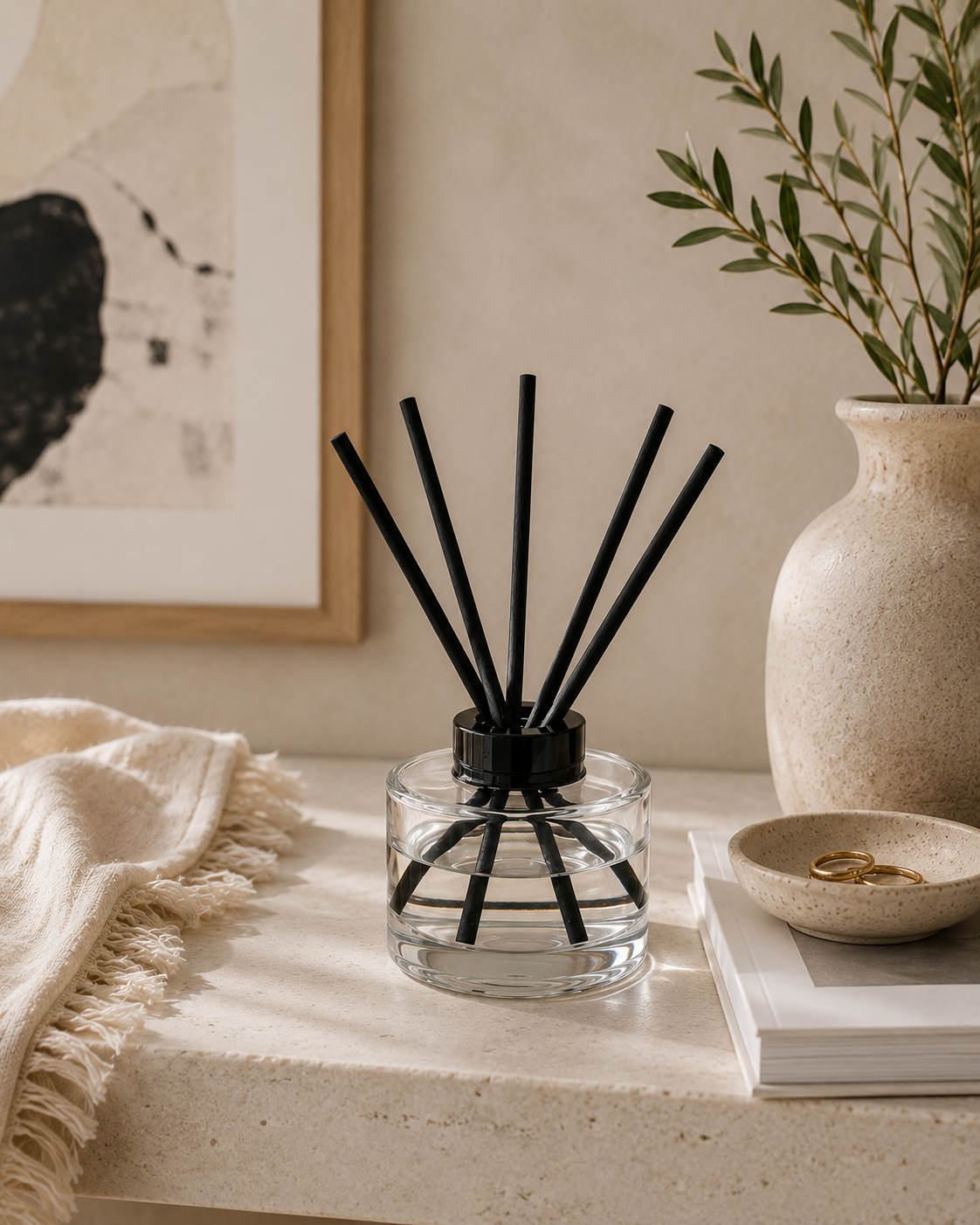 A clear glass reed diffuser bottle with black reeds and a black collar, styled on a beige stone surface with a cream throw blanket, framed wall art, a ceramic vase with greenery, and a small dish.
