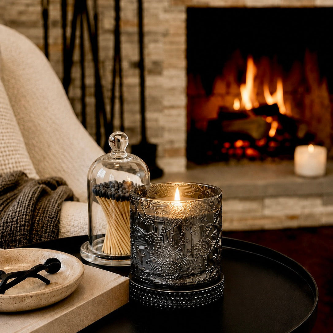 A lit black textured glass candle sits on a black round table beside a glass cloche with matches and a small tray in a cozy living room setting with a fireplace in the background.