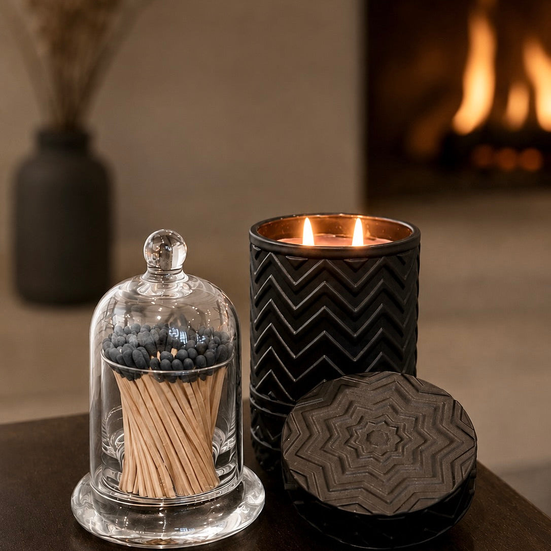 A styled home decor scene featuring a black candle tin with a chevron pattern and lit flames, a glass cloche containing wooden matches and blue berries, and a round lid with a geometric mandala pattern, set on a dark wooden table with a blurred fireplace in the background.