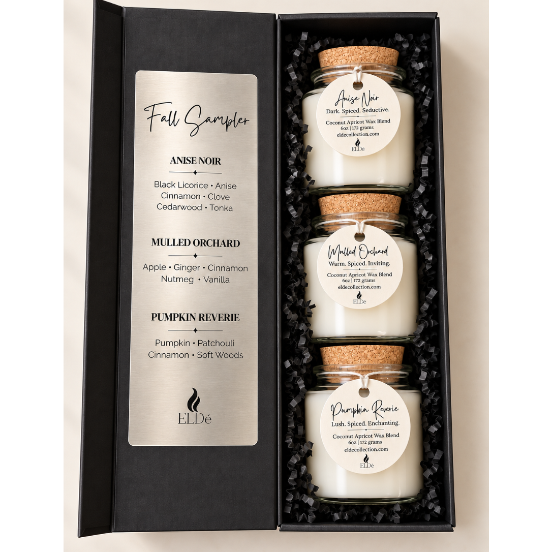 A gift set of three ELDE candles in glass jars with cork lids, presented inside an open black box with a printed scent list labeled 'Fall Sampler.' The candles are arranged in black shredded paper, and the labels show different spice and autumn-themed scents: Anise Noir, Mulled Orchard, and Pumpkin Reverie.