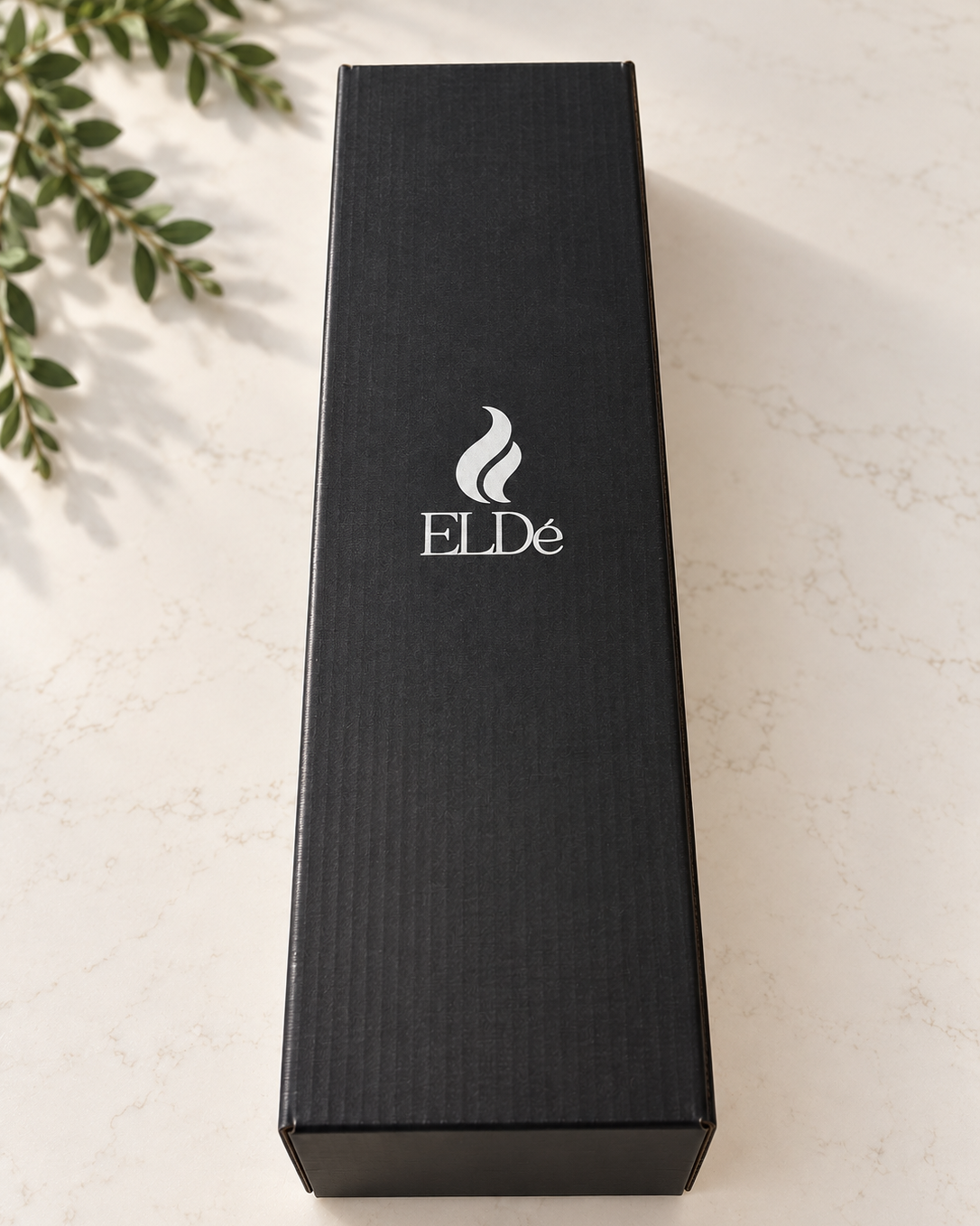 A tall, black rectangular product box with the brand name 'ELDé' and a stylized flame logo printed in white on the front, placed on a light marble surface with green leaves in the corner.