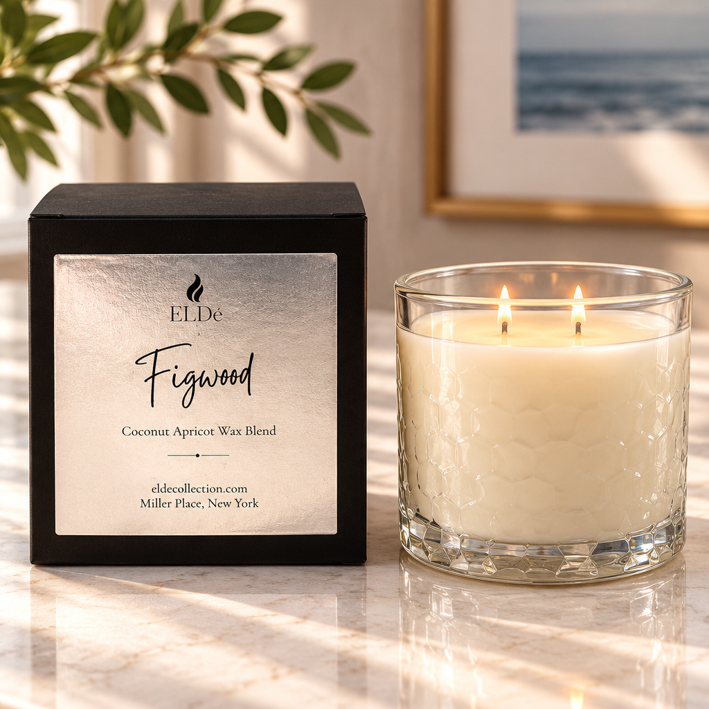A lit double-wick scented candle in a textured clear glass jar sits beside its black branded box on a marble surface. The label reads ELDE Figwood and shows coconut apricot wax blend.
