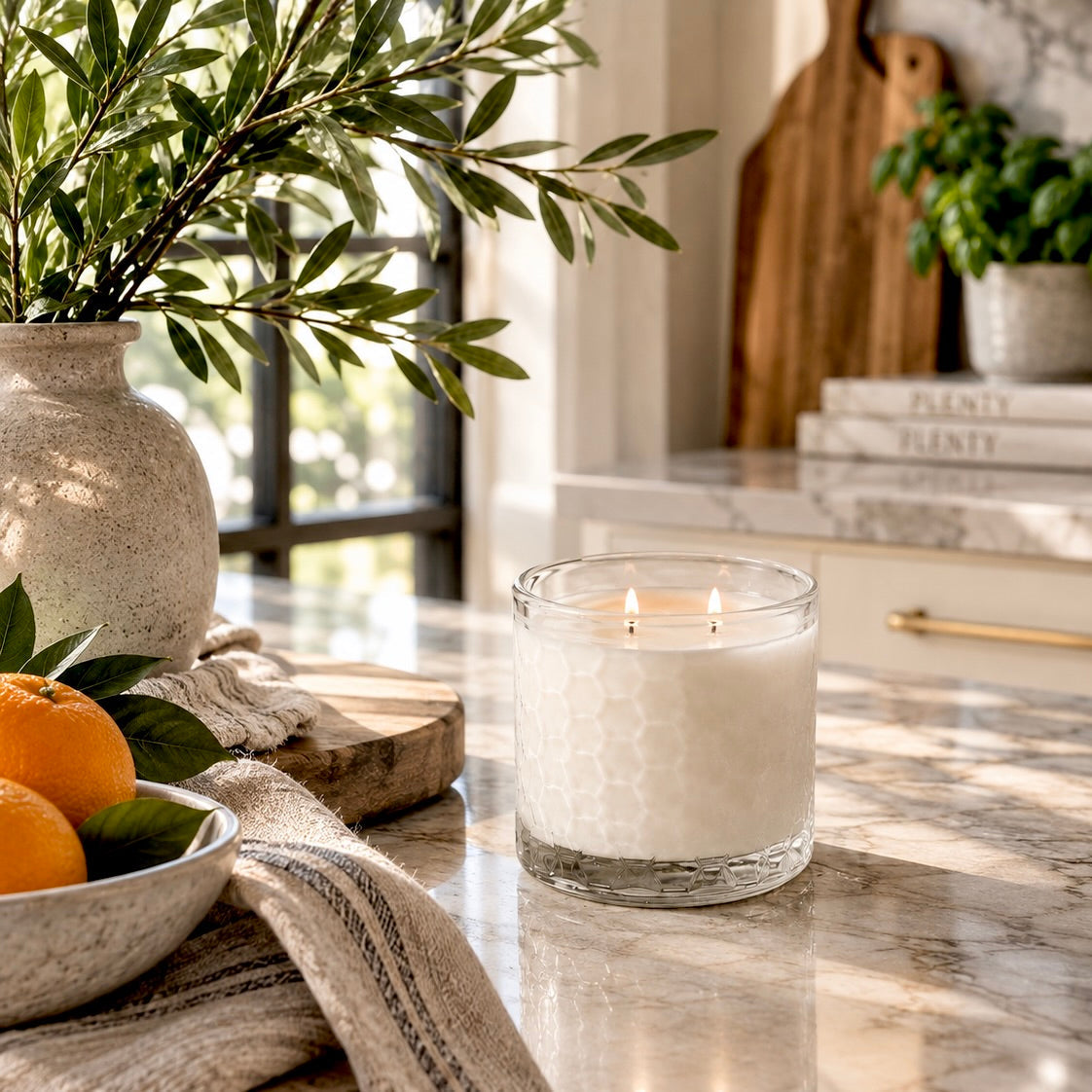 A white wax candle in a clear glass jar with a textured honeycomb pattern, lit with two flames, set on a marble countertop in a bright, modern kitchen.
