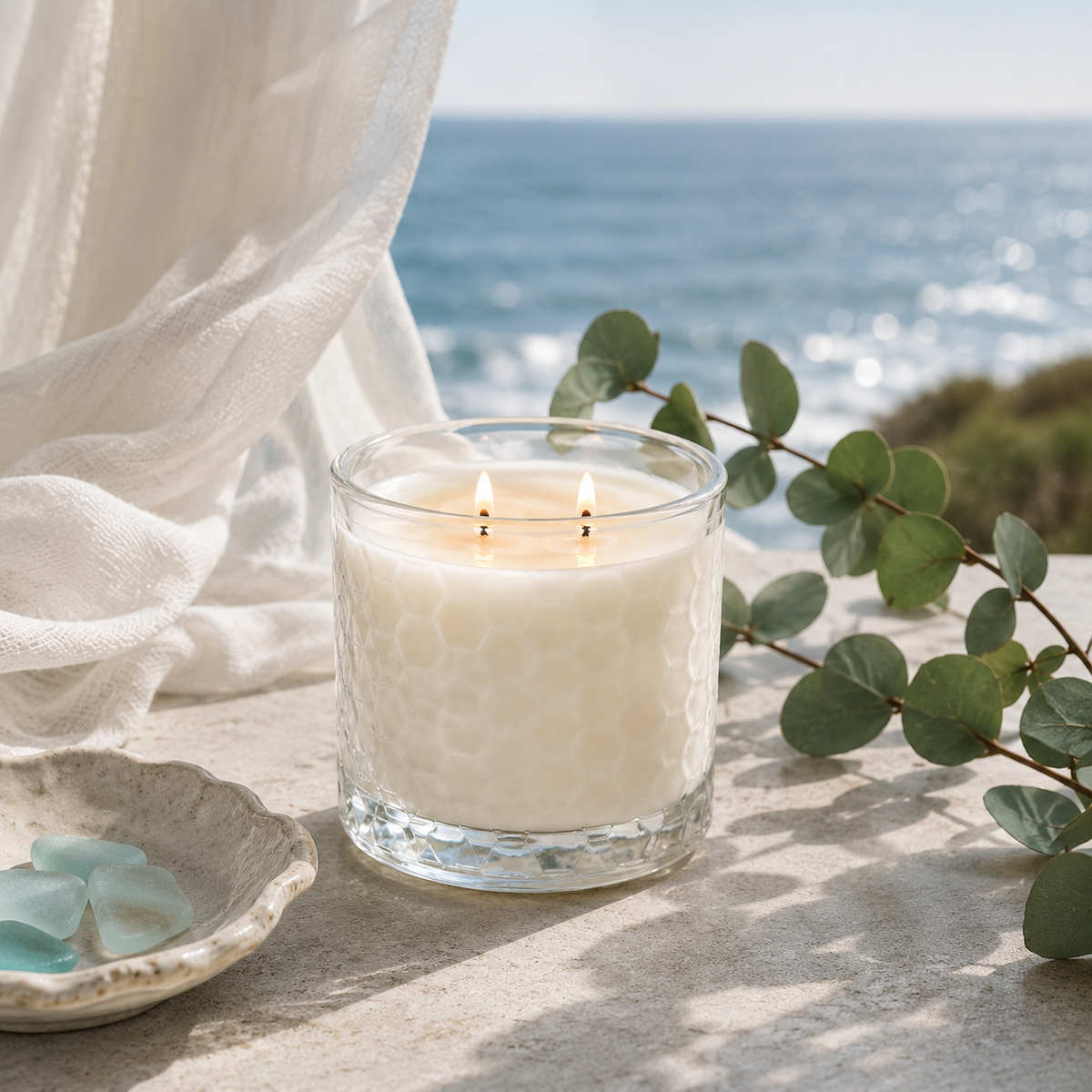 A lit double-wick candle in a textured clear glass jar sits on a stone surface outdoors by the ocean, with white draped fabric, green eucalyptus leaves, and a small dish of blue stones nearby.